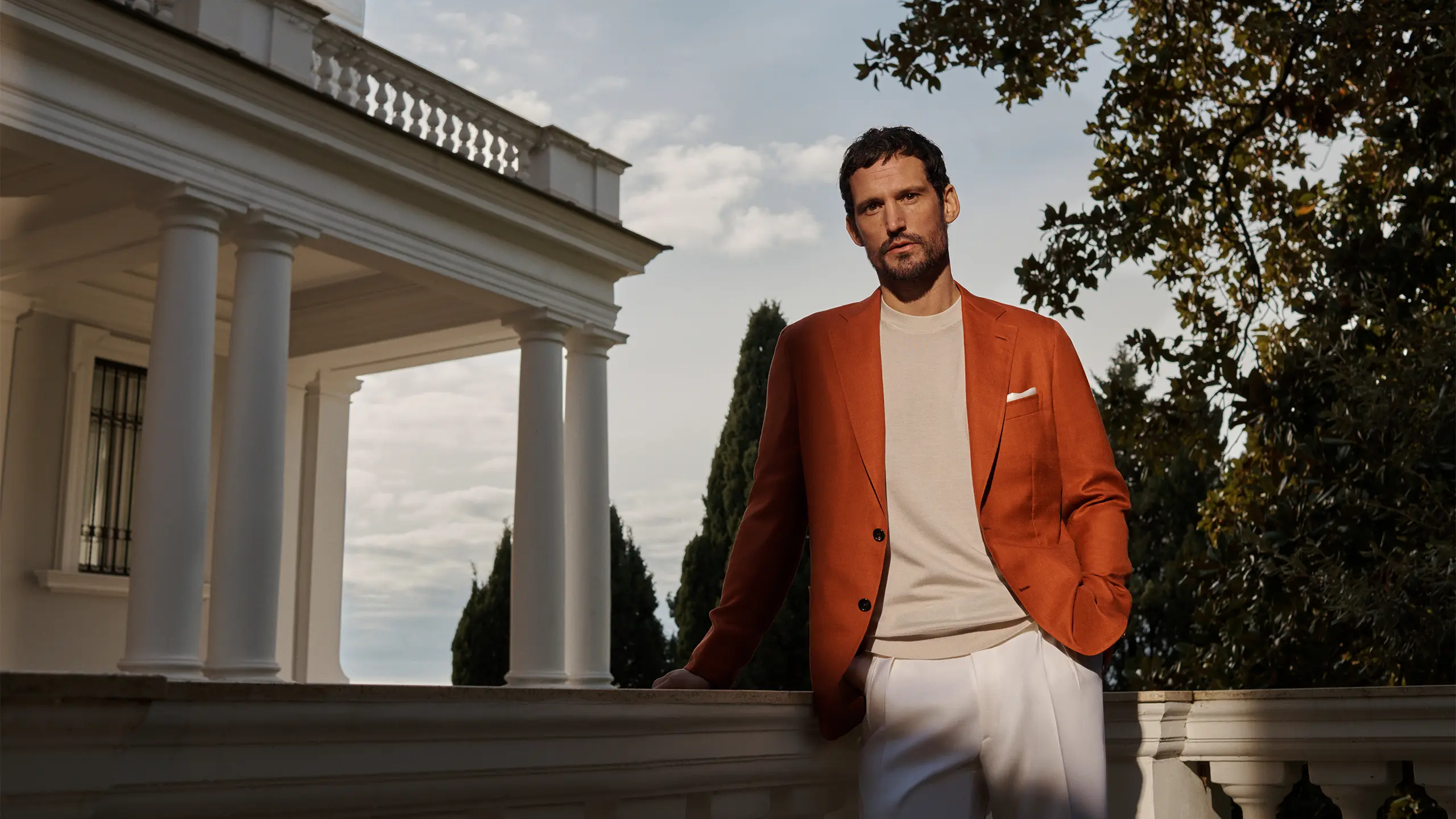 Man in Brioni rust-orange blazer and cream shirt stands outdoors near white columned building with trees in background.