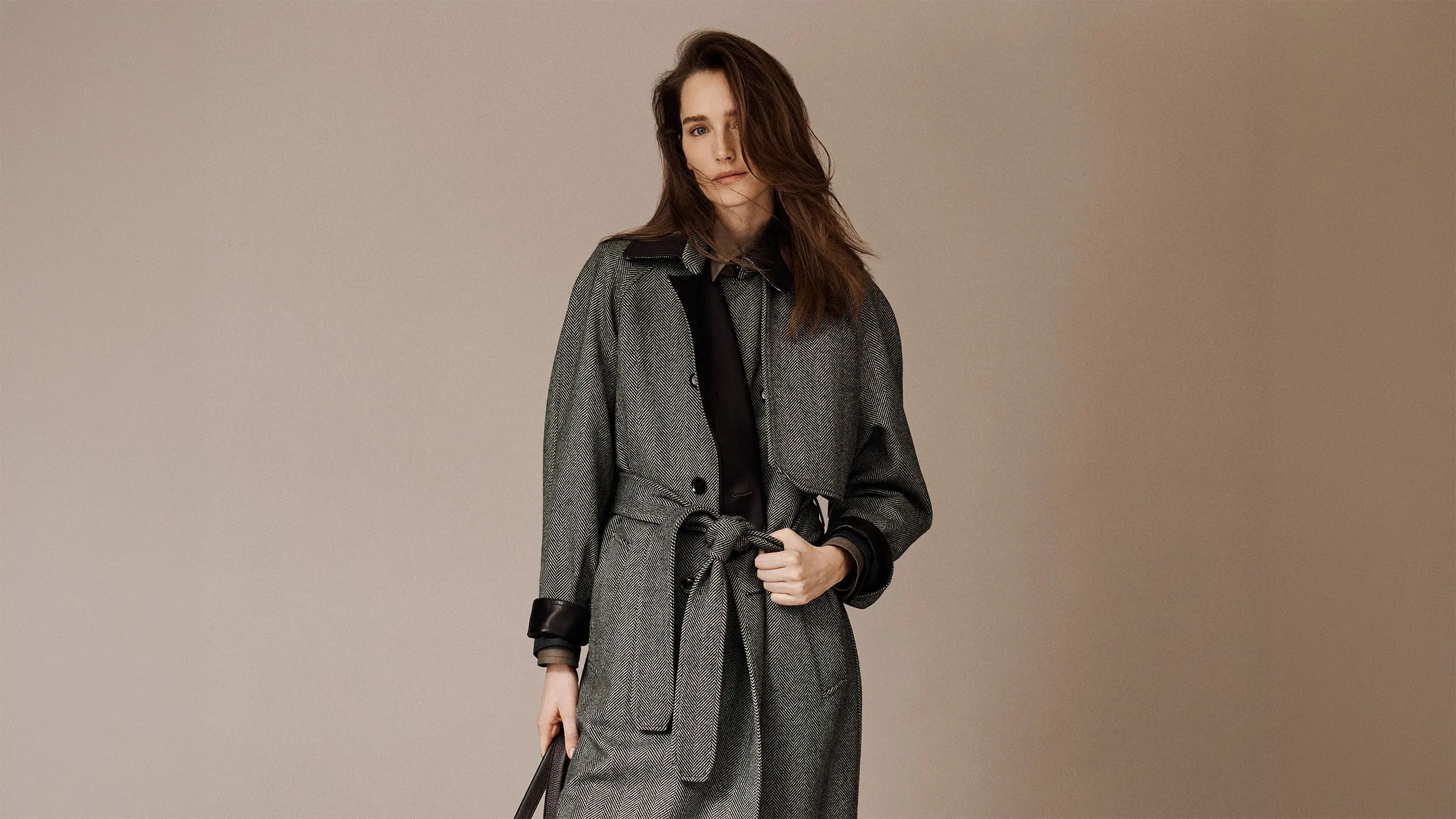 Woman with long brown hair wearing a Brioni gray herringbone coat with black trim and belt against a beige background.