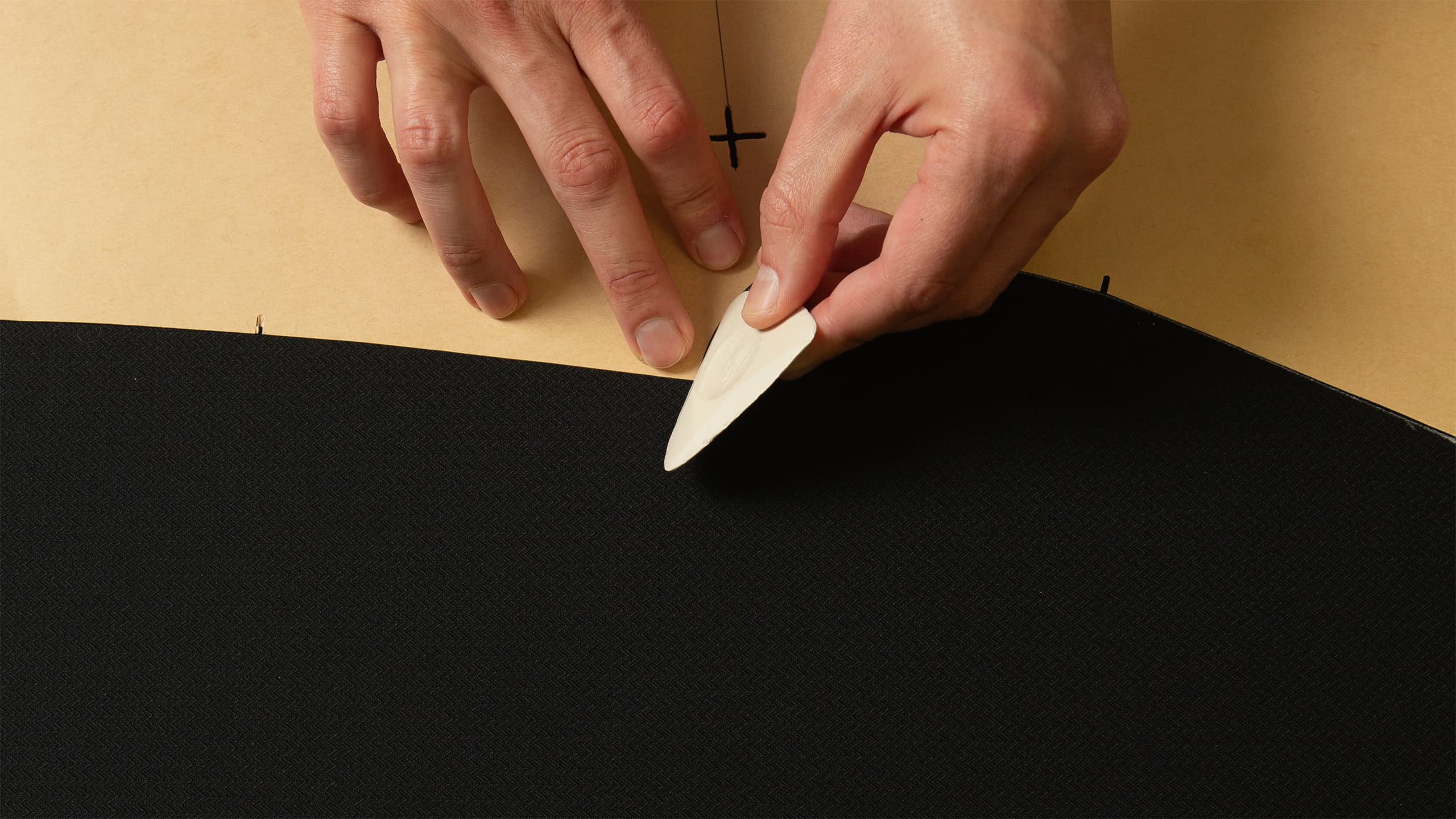 Hands using a bone folder tool to crease paper, working with black and tan materials for crafting or bookbinding.