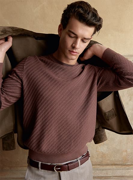 Model in Trama Brioni brown sweater putting on a beige jacket, wearing light pants with a brown leather belt.