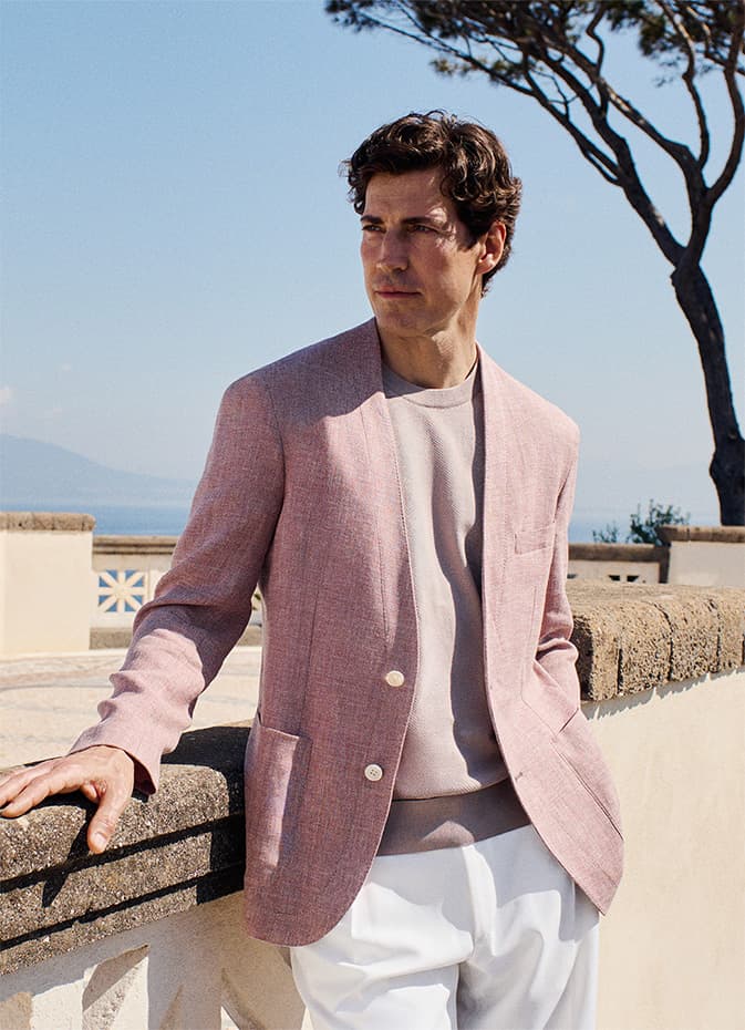 Man in Brioni pink linen blazer and white pants standing on Mediterranean terrace with tree and blue sky in background.
