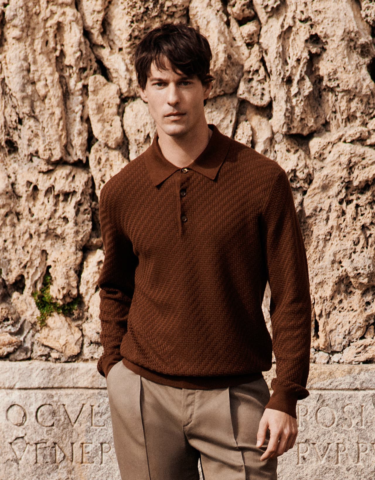Man in Brioni brown textured polo shirt and beige trousers standing against weathered stone wall with carved text.