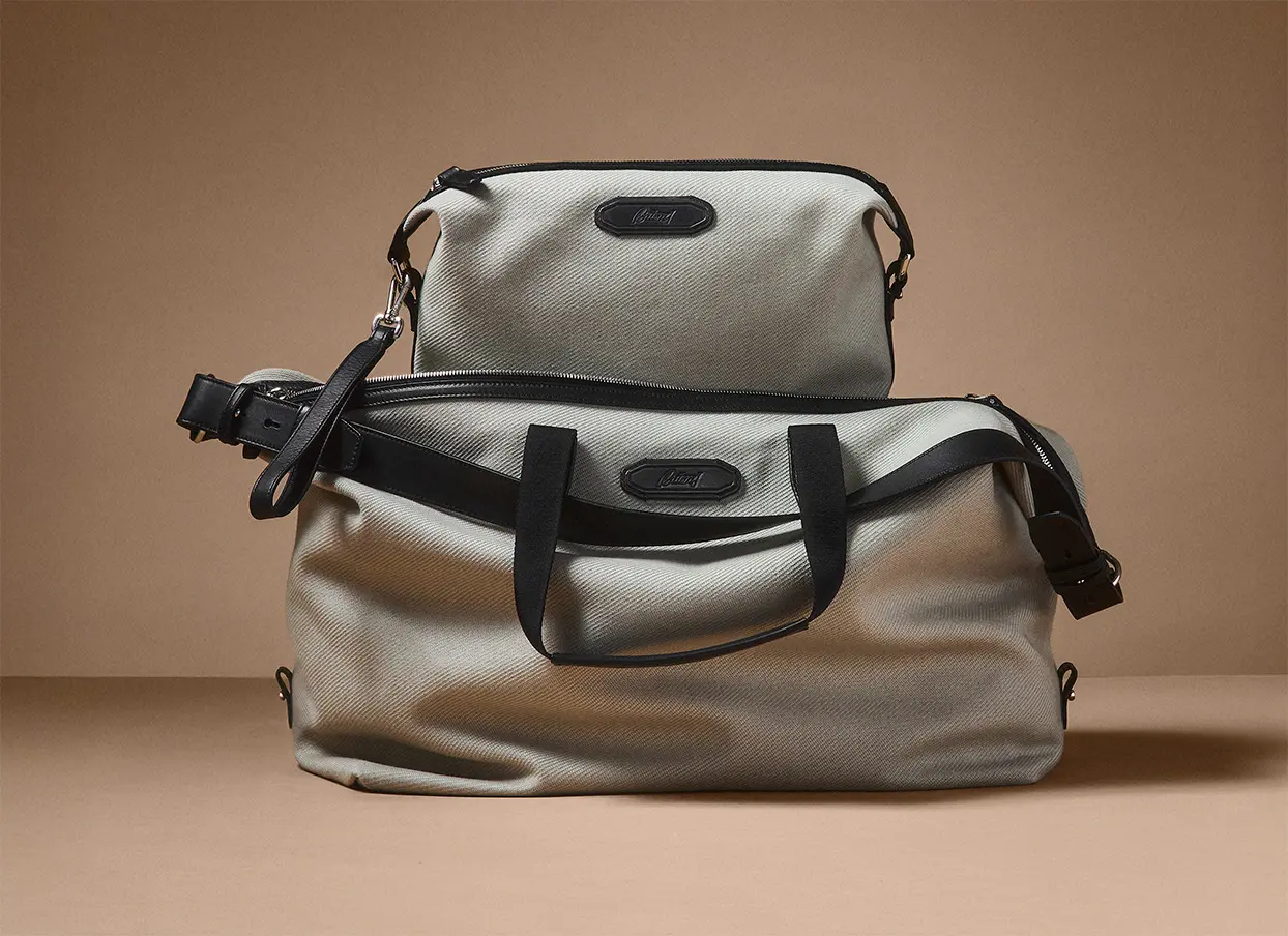 Two Brioni light grey travel bags with black trim and handles displayed against a neutral beige background.