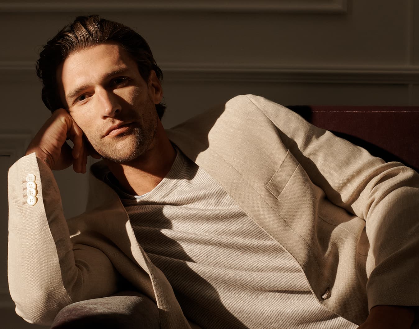 Man in Brioni beige blazer and striped shirt reclining in warm sunlight against neutral wall, hand touching his head.