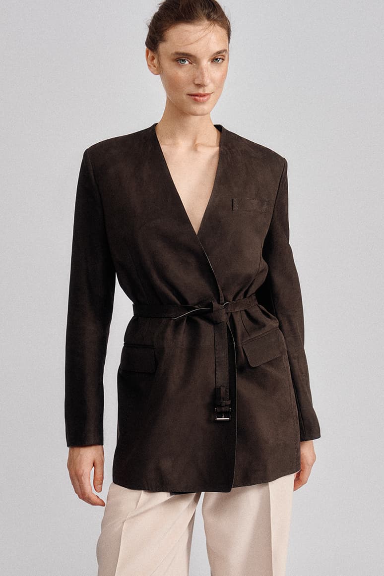 Woman wearing Brioni dark brown suede wrap jacket with tie belt and chest pocket, paired with cream pants on gray background.