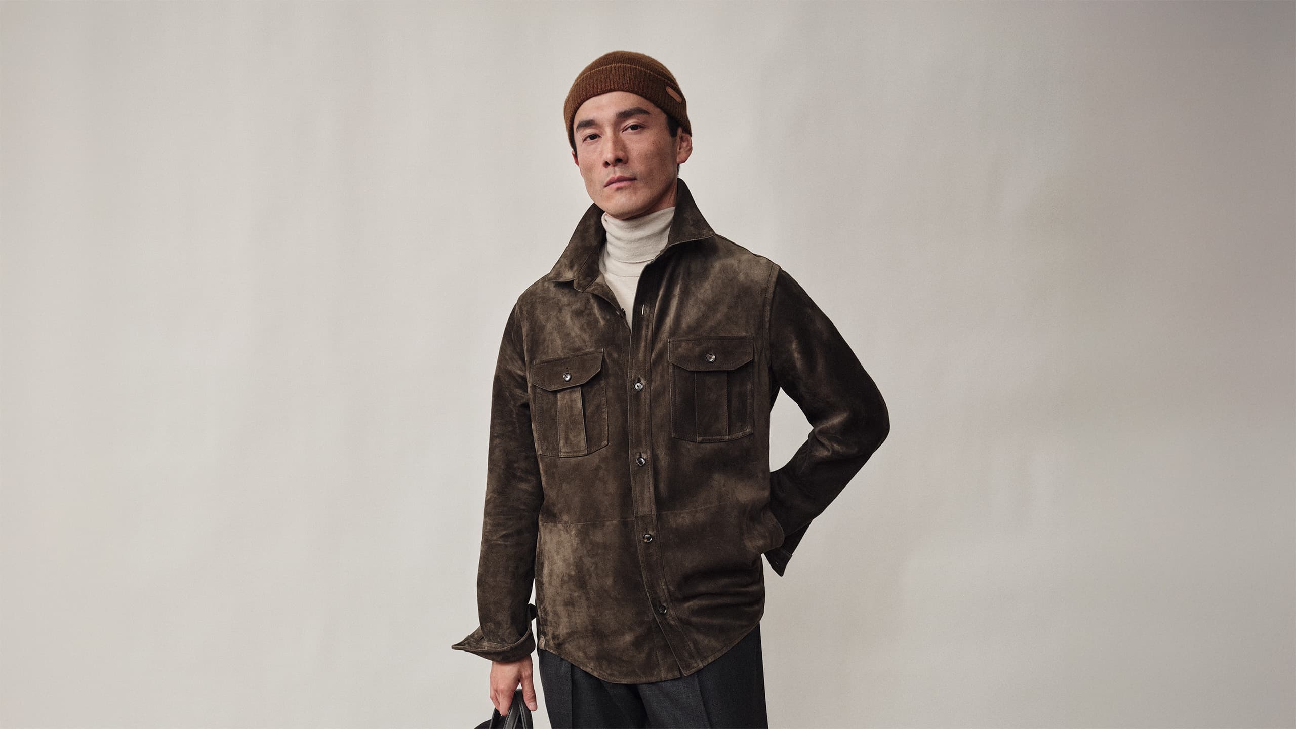 Person wearing Brioni brown suede jacket, white turtleneck, and brown beanie against neutral background.