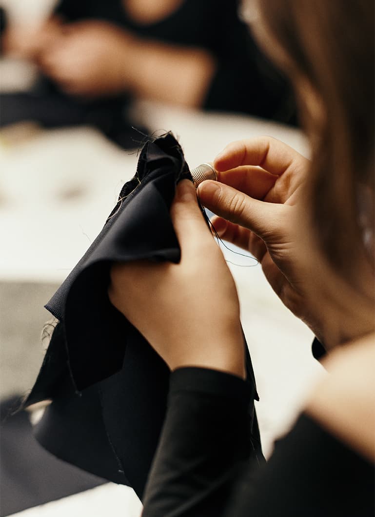 Primo piano delle mani che cuciono un tessuto nero con un ditale, mostrando l’accuratezza della lavorazione artigianale in un laboratorio di moda.