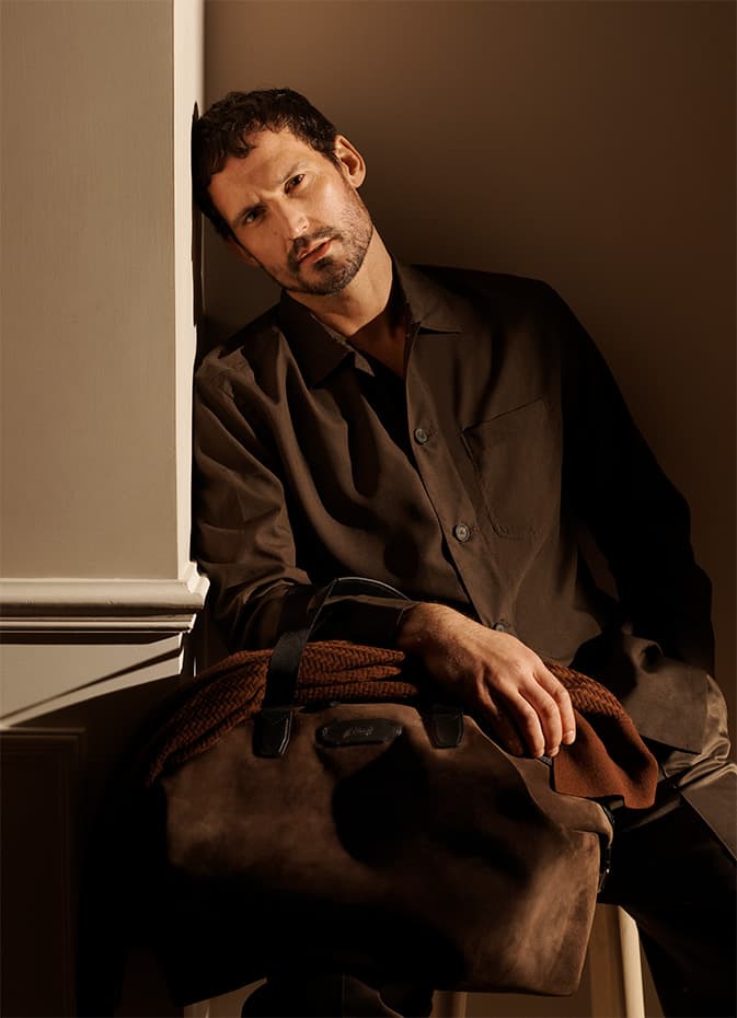 Man in Brioni brown shirt leaning against white wall in warm lighting, holding rust-colored sweater, gazing at camera pensively.
