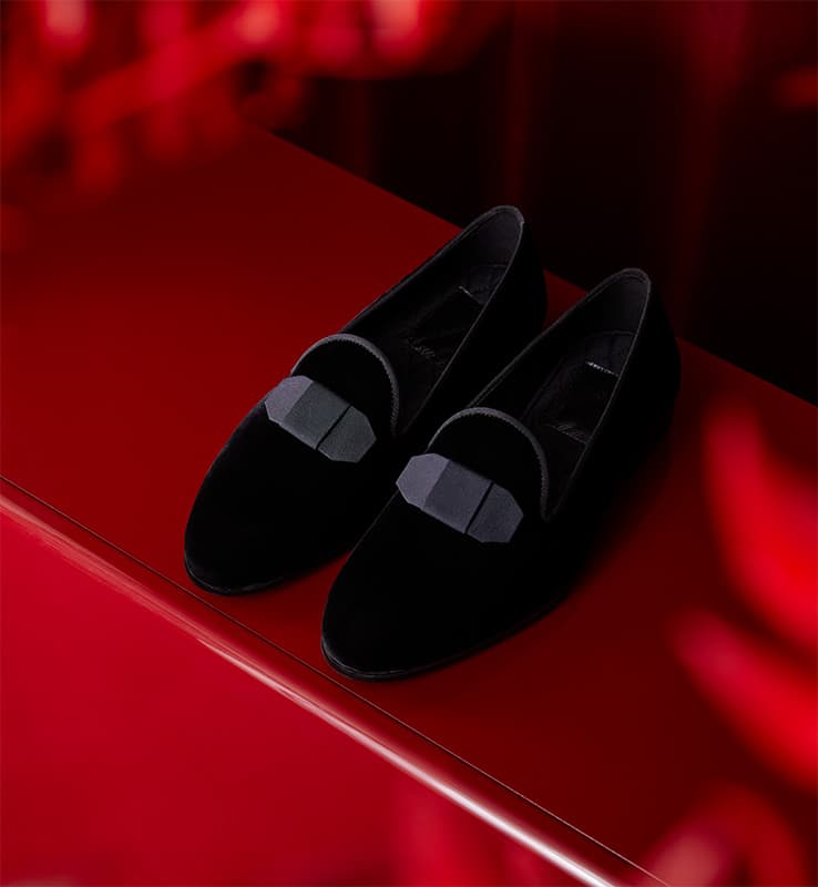 Elegant Brioni black velvet loafers with bow details displayed on a glossy red surface against a dramatic red background.