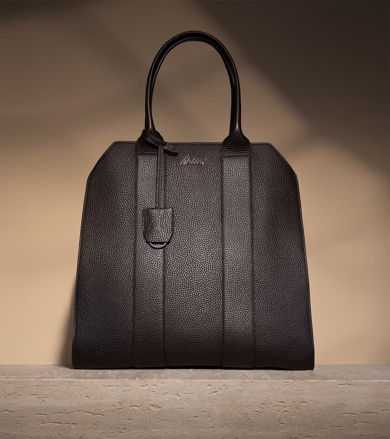 Brioni Black textured leather handbag with double handles and brand logo, standing upright against a beige gradient background.
