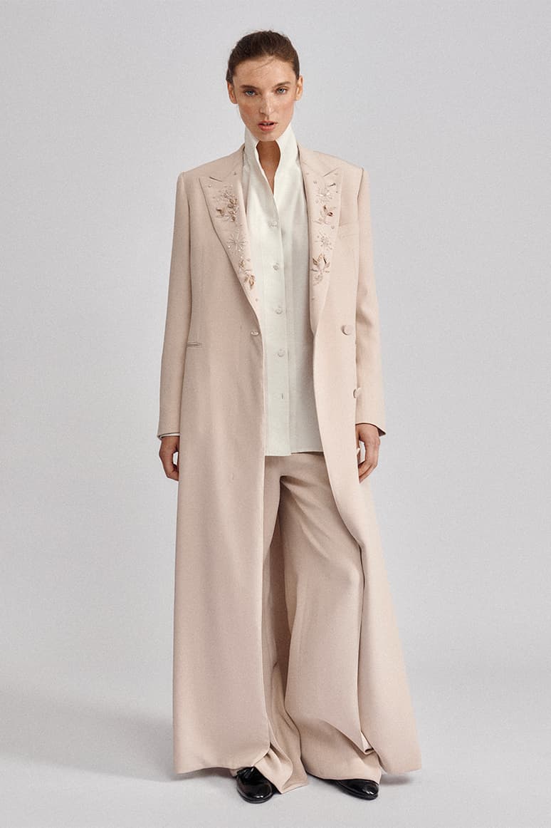 Woman in Brioni beige embroidered long coat over white blouse and wide-leg pants against gray background.
