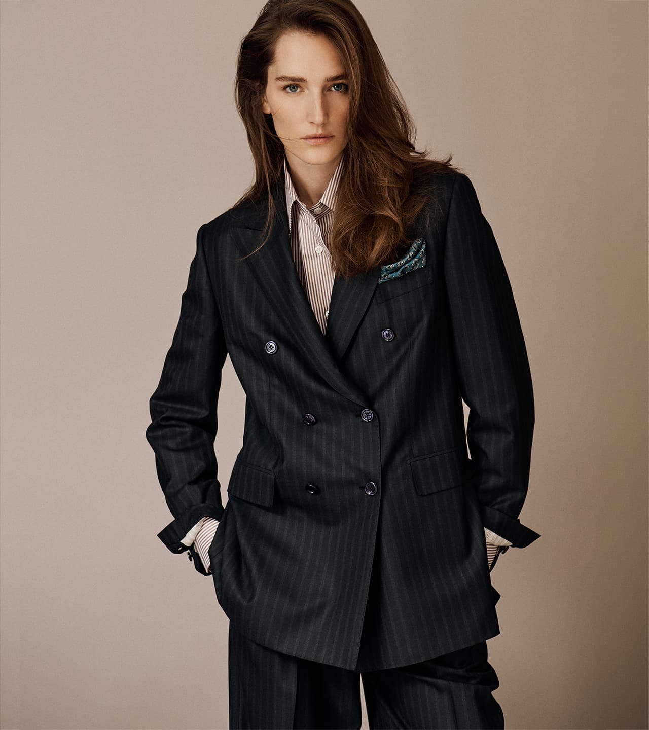 Woman with long brown hair wearing Brioni dark pinstripe double-breasted blazer over striped shirt against beige background.