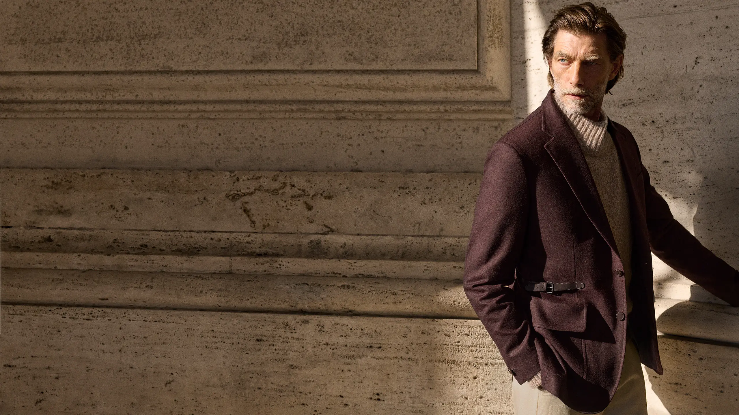 Distinguished man in Brioni burgundy blazer and beige turtleneck standing against textured concrete wall with natural lighting.