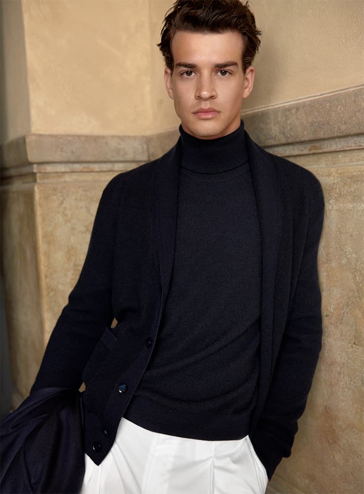 Person in elegant Brioni navy turtleneck and cardigan with white pants against beige stone wall background.