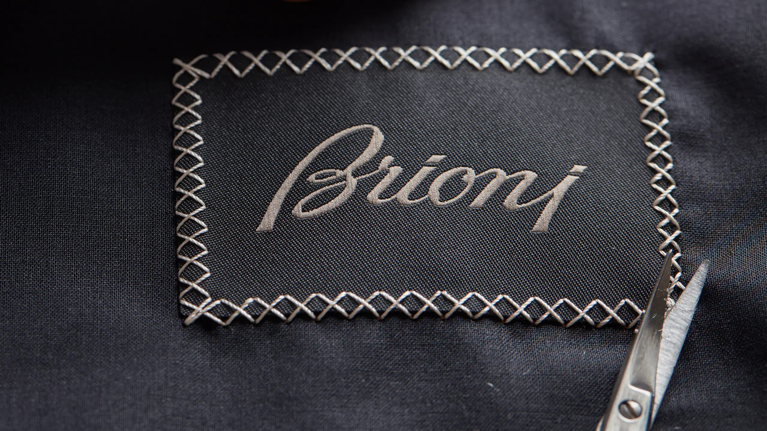 Elegant "Brioni" logo embroidered in beige thread on dark fabric with decorative cross-stitch border and scissors visible.
