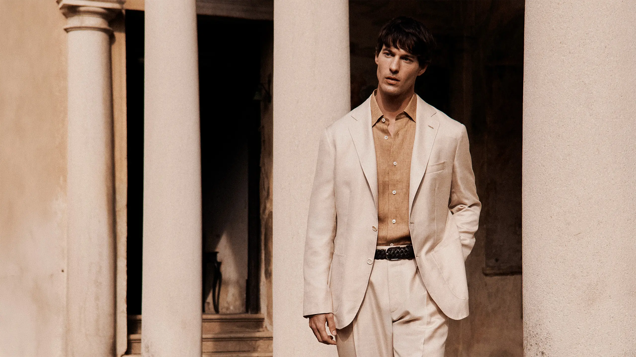 Young man in Brioni light blue blazer and beige pants standing against weathered beige and brown textured wall with natural lighting.