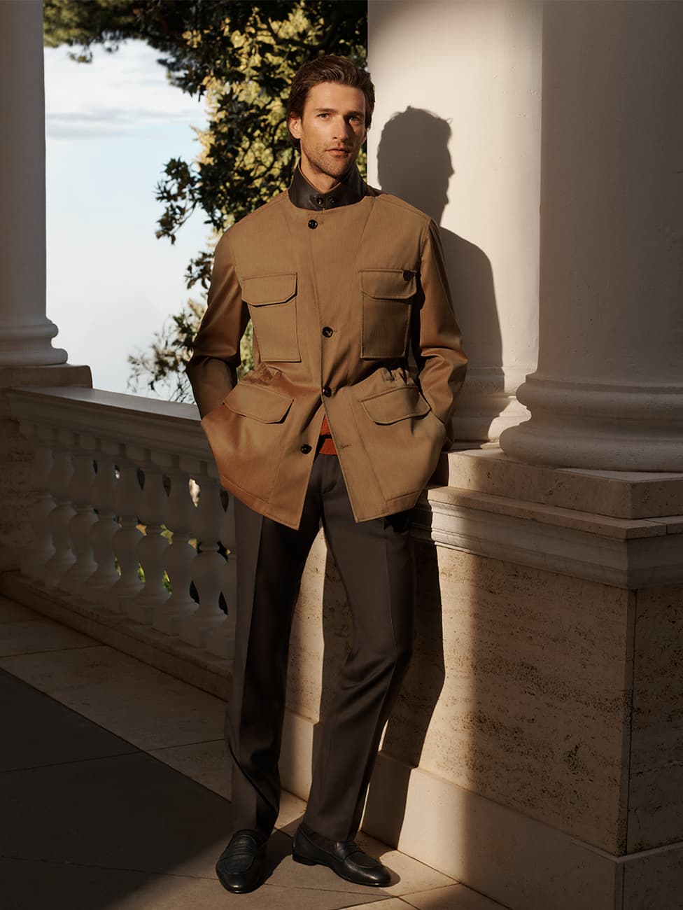 Man in tan Brioni utility jacket with multiple pockets and black pants standing on terrace with white columns and ocean view.