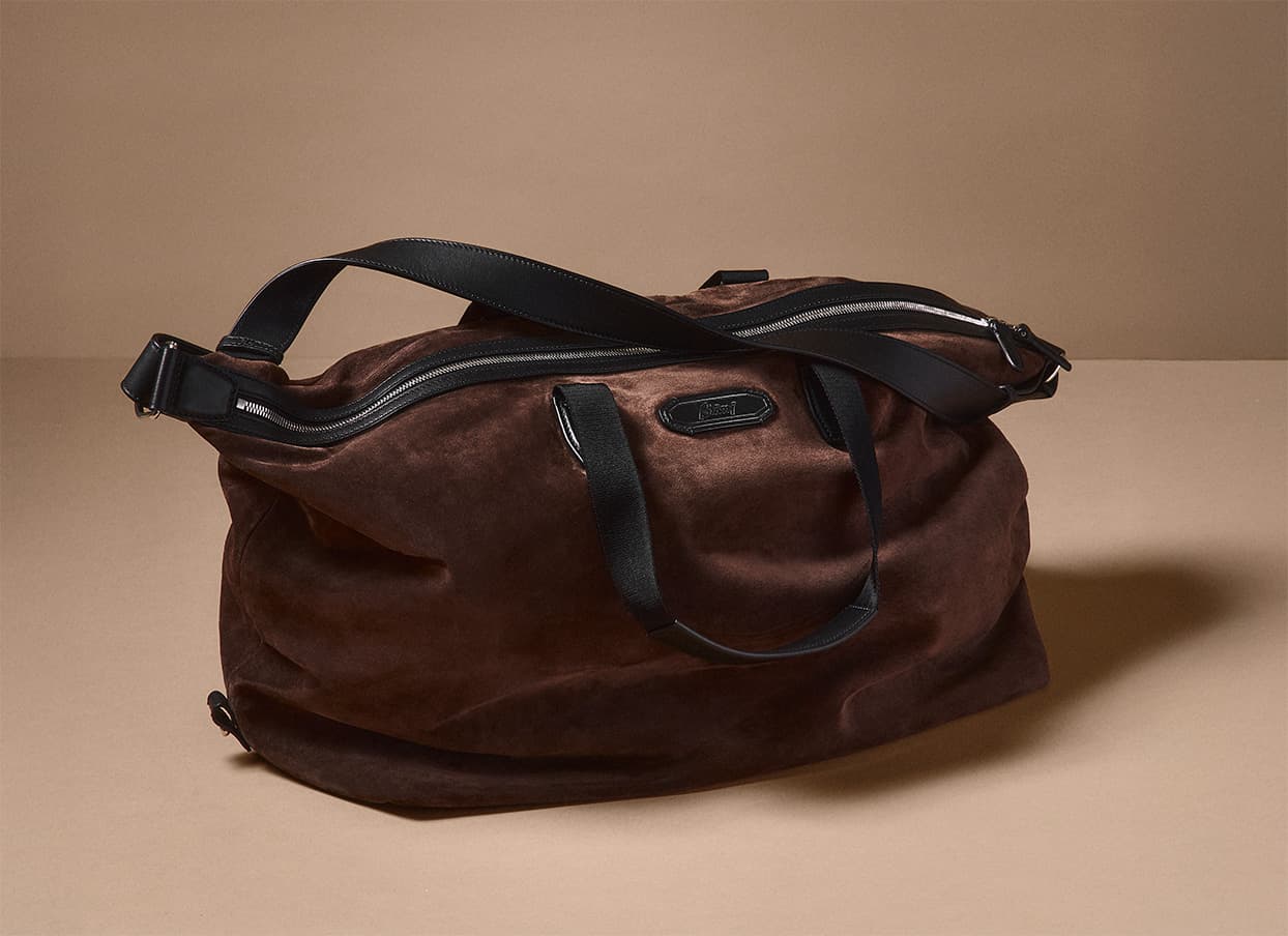 Brioni brown suede weekender bag with black leather straps and zipper details against a neutral beige background.