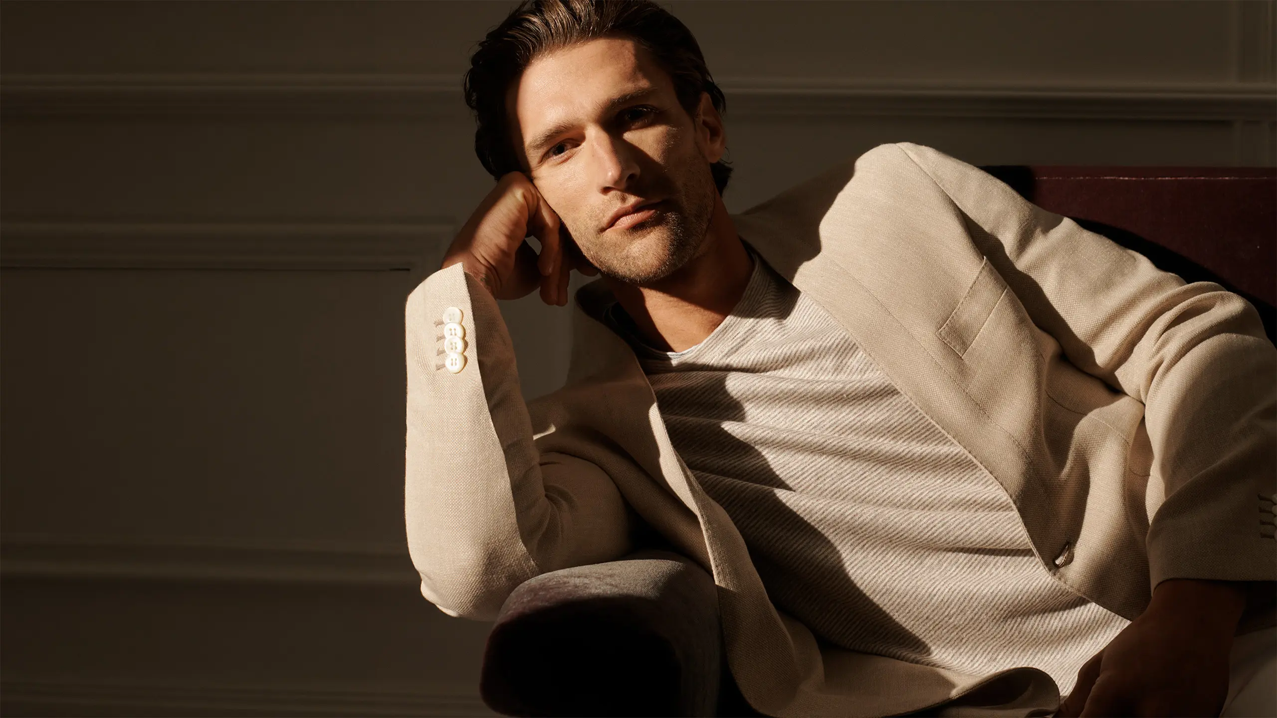 Man in Brioni beige blazer and white shirt reclining in warm sunlight against neutral wall, hand near face in relaxed pose.