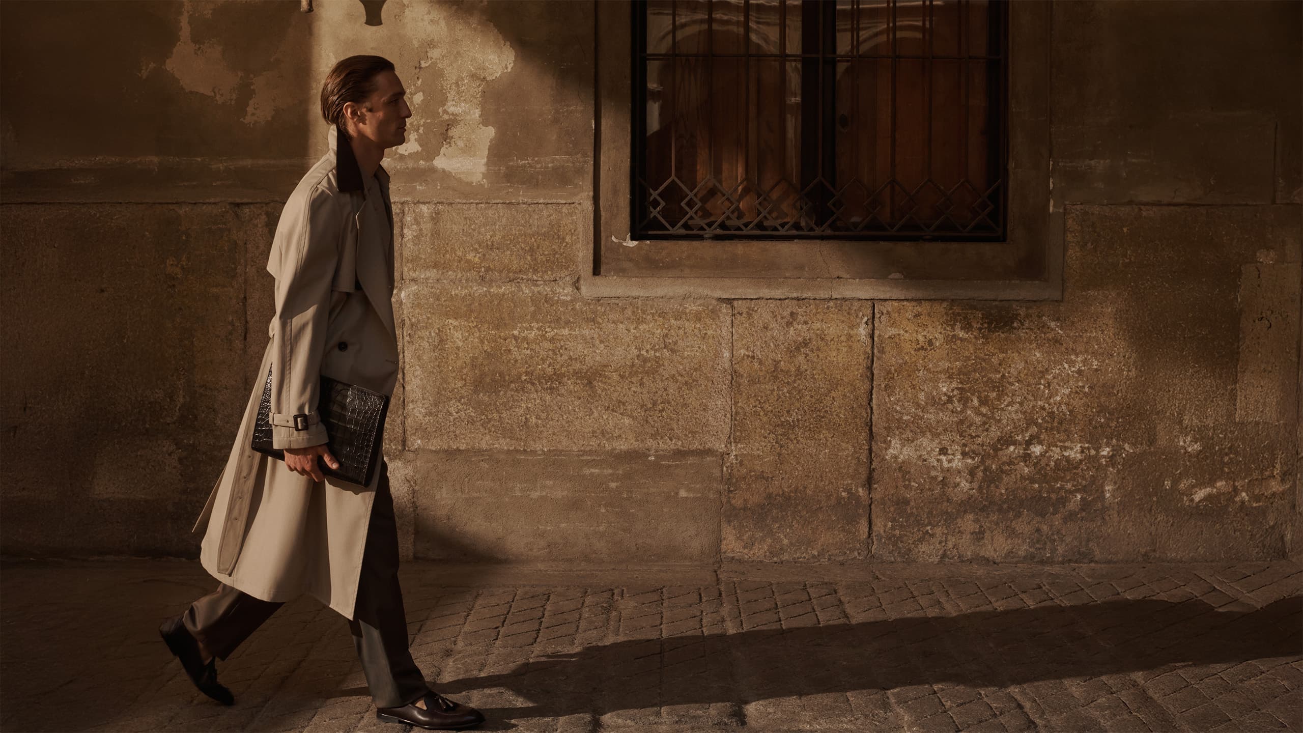 Man in beige trench coat walking past weathered stone wall with barred window, carrying dark clutch bag in warm sunlight.