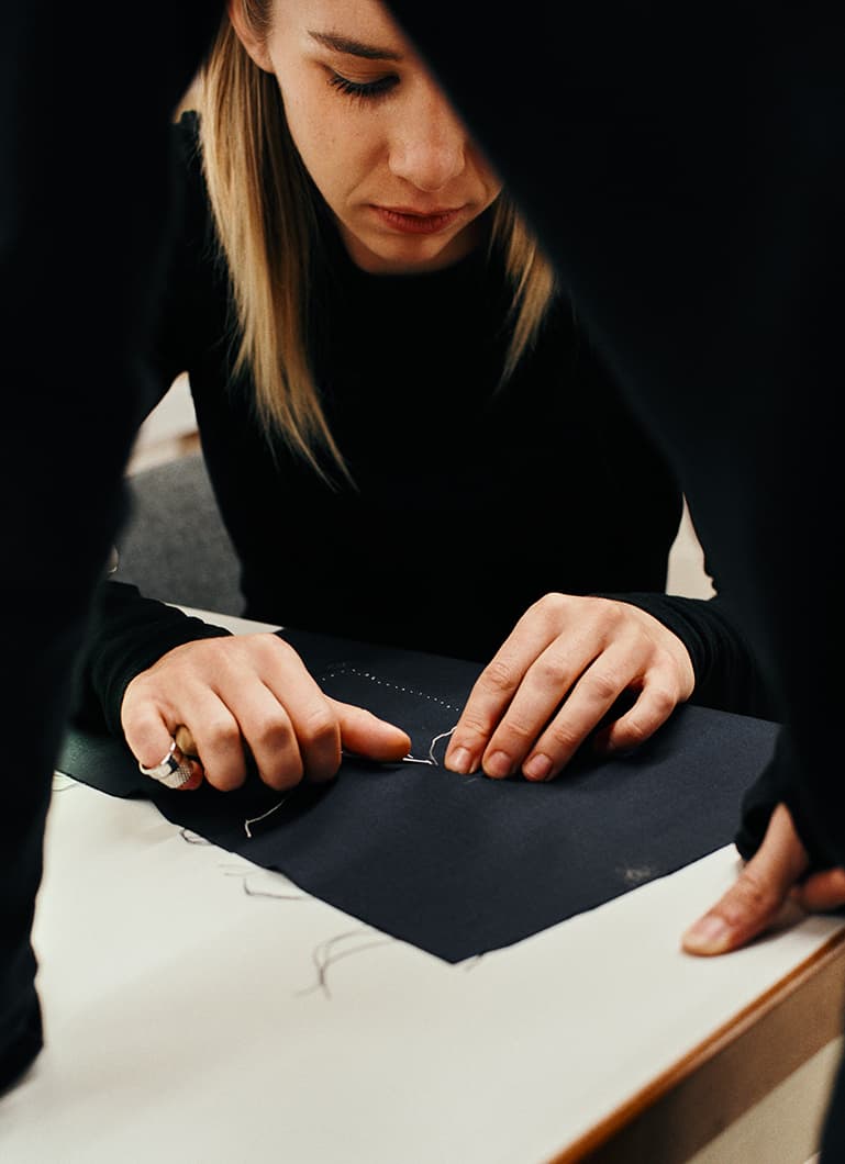 Una giovane studentessa è ritratta mentre cuce con cura un tessuto scuro, concentrata su un lavoro artigianale dettagliato in una luce soffusa.