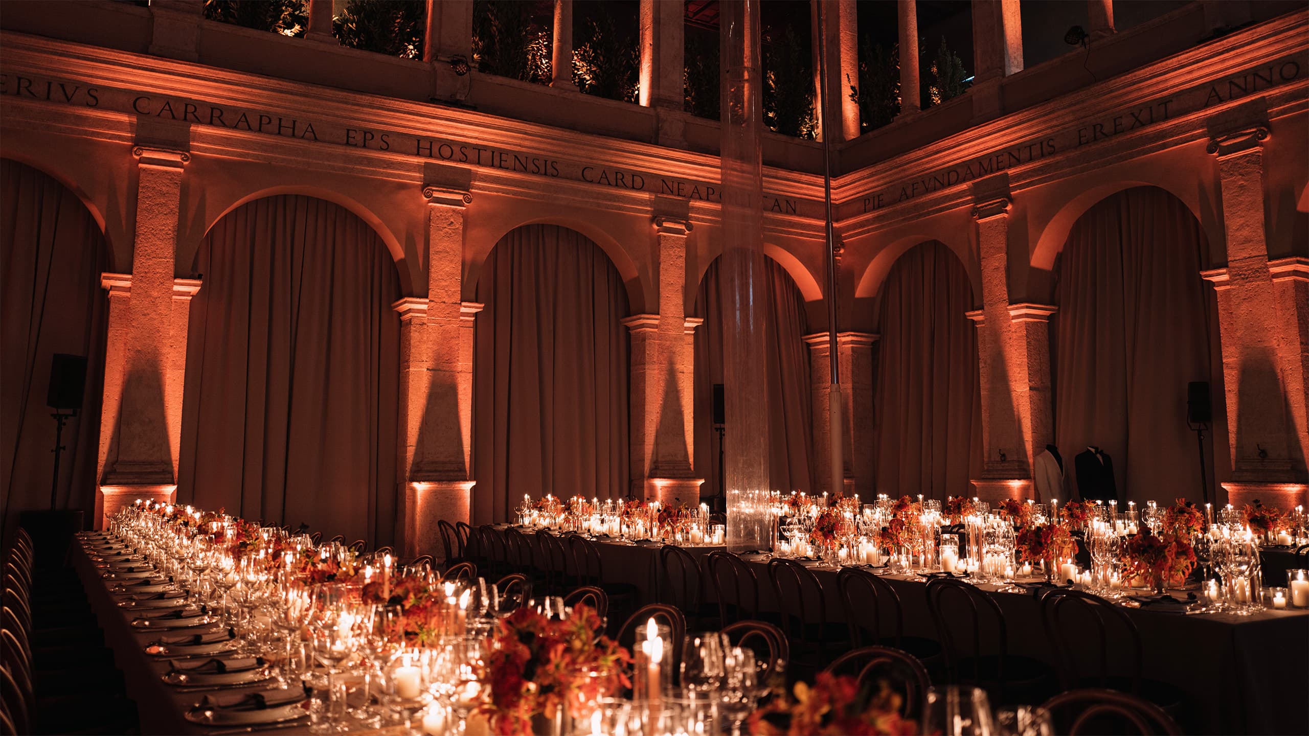 Elegant banquet tables with candles and floral arrangements in a historic venue with arched walls bathed in amber light.