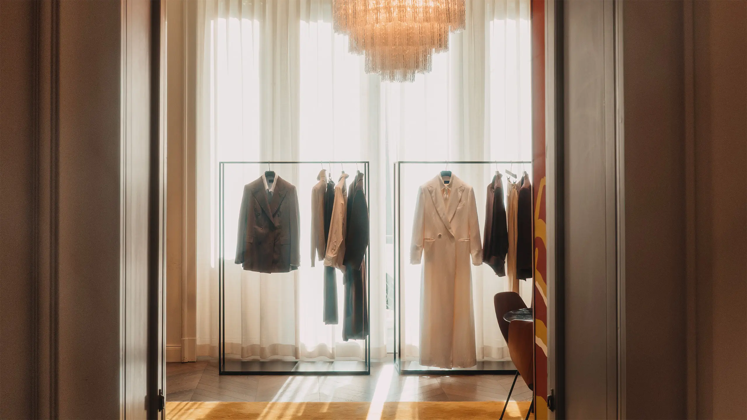 Elegant glass wardrobes displaying Brioni suits and coats beneath a golden chandelier in a luxury boutique with white curtains.