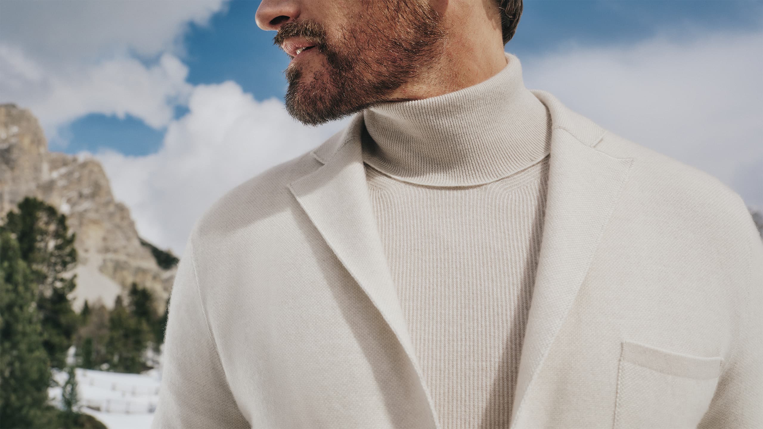 Person wearing Brioni cream turtleneck and blazer against snowy mountain backdrop with blue sky and evergreen trees.