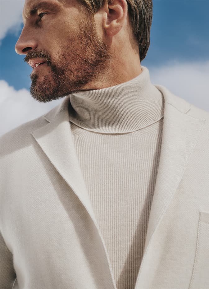Profile view of person with beard wearing Brioni cream turtleneck sweater and light blazer against blue sky background.