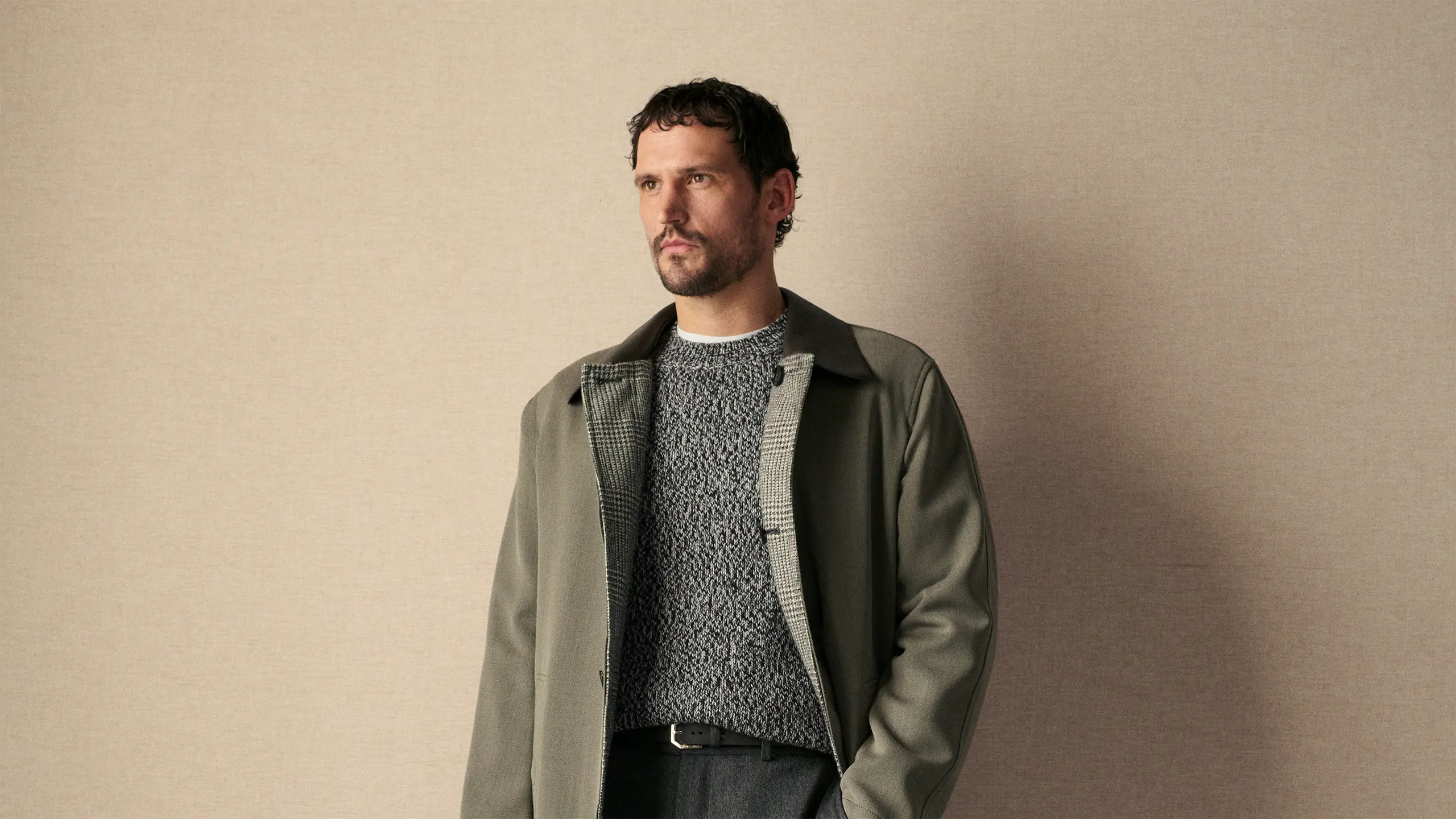 Man with dark hair and beard wearing Brioni olive green jacket over gray knit sweater against beige background, looking upward.