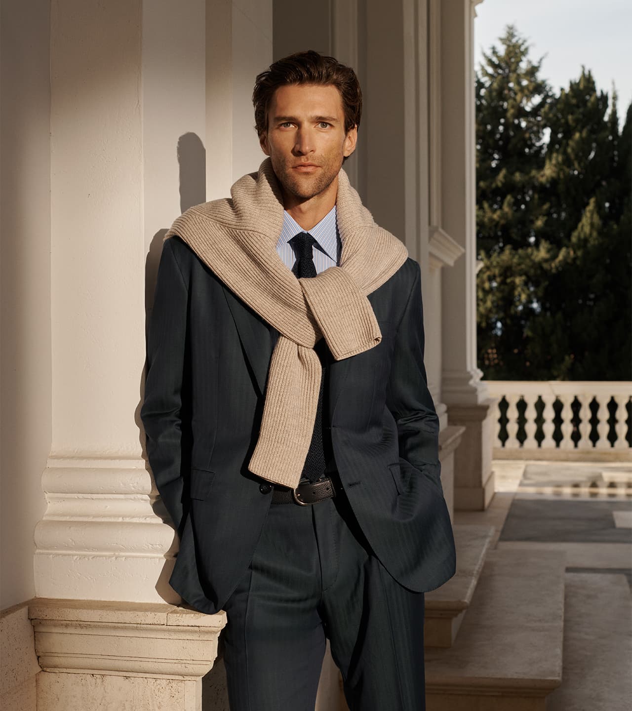 Man in Brioni dark green suit with beige scarf and blue striped shirt standing by white columns with evergreen trees in background