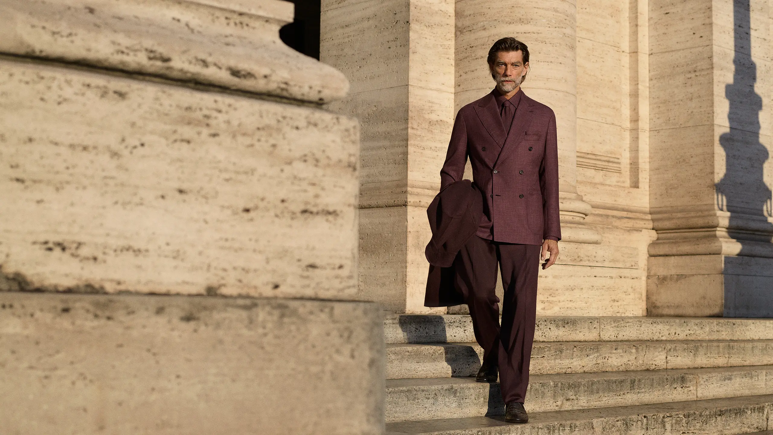 Man in Brioni burgundy double-breasted suit standing on stone steps between classical columns, holding a coat over his arm.