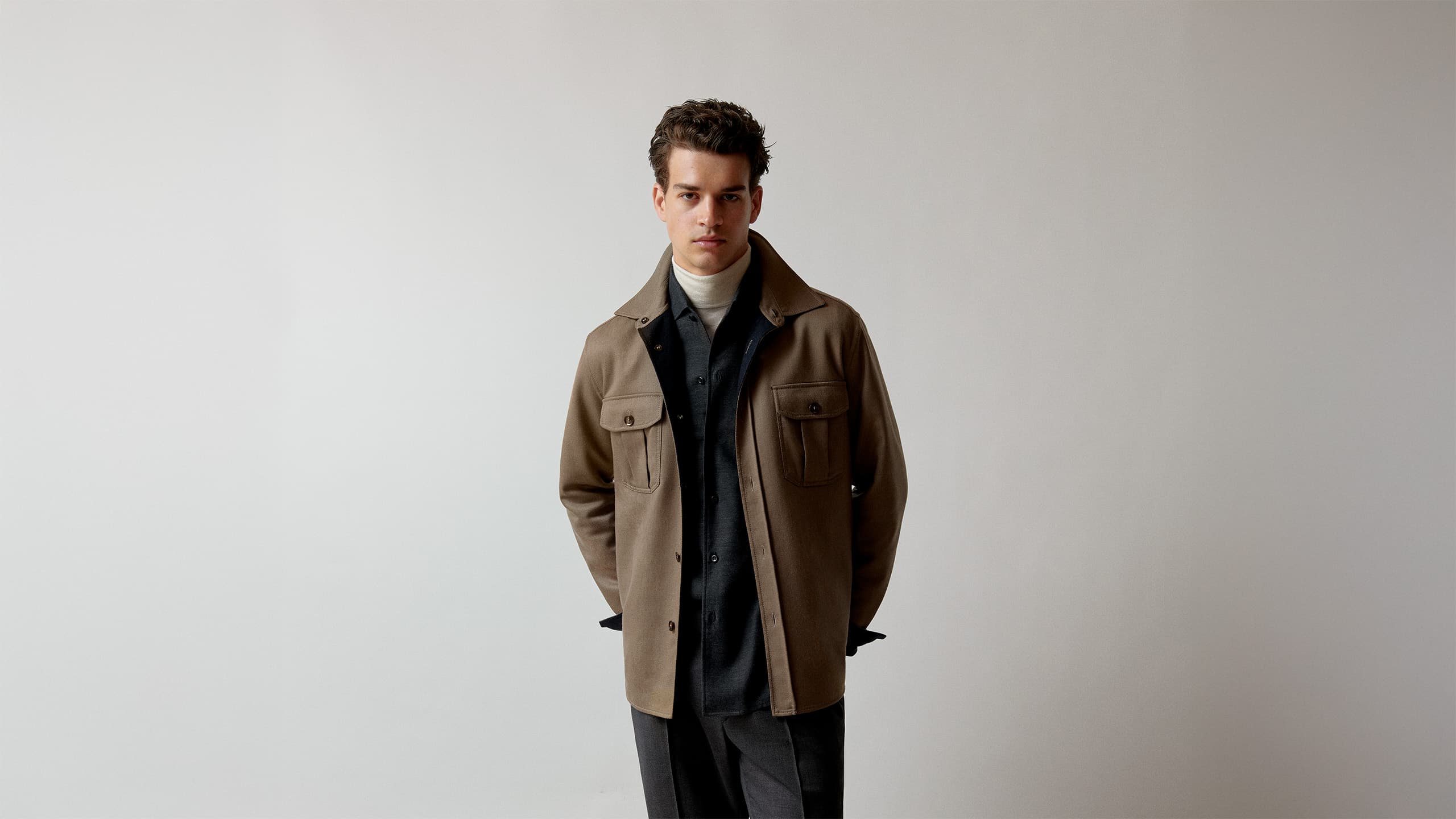 Person wearing layered autumn outfit with beige Brioni jacket over dark shirt and white turtleneck against neutral background.