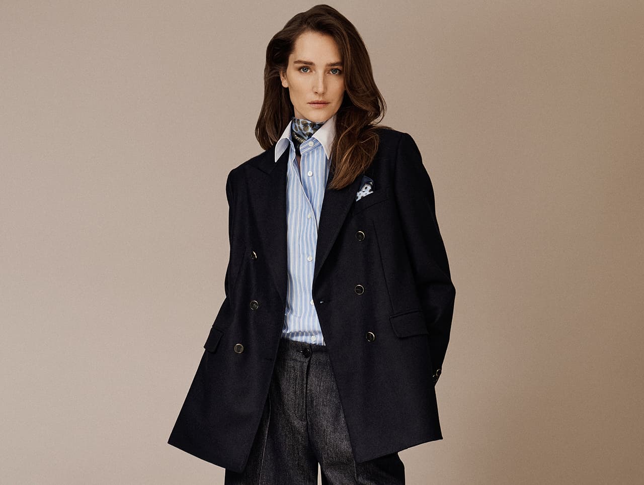 Woman in Brioni gray tailored suit sitting against textured brown wall, looking directly at camera with serious expression.