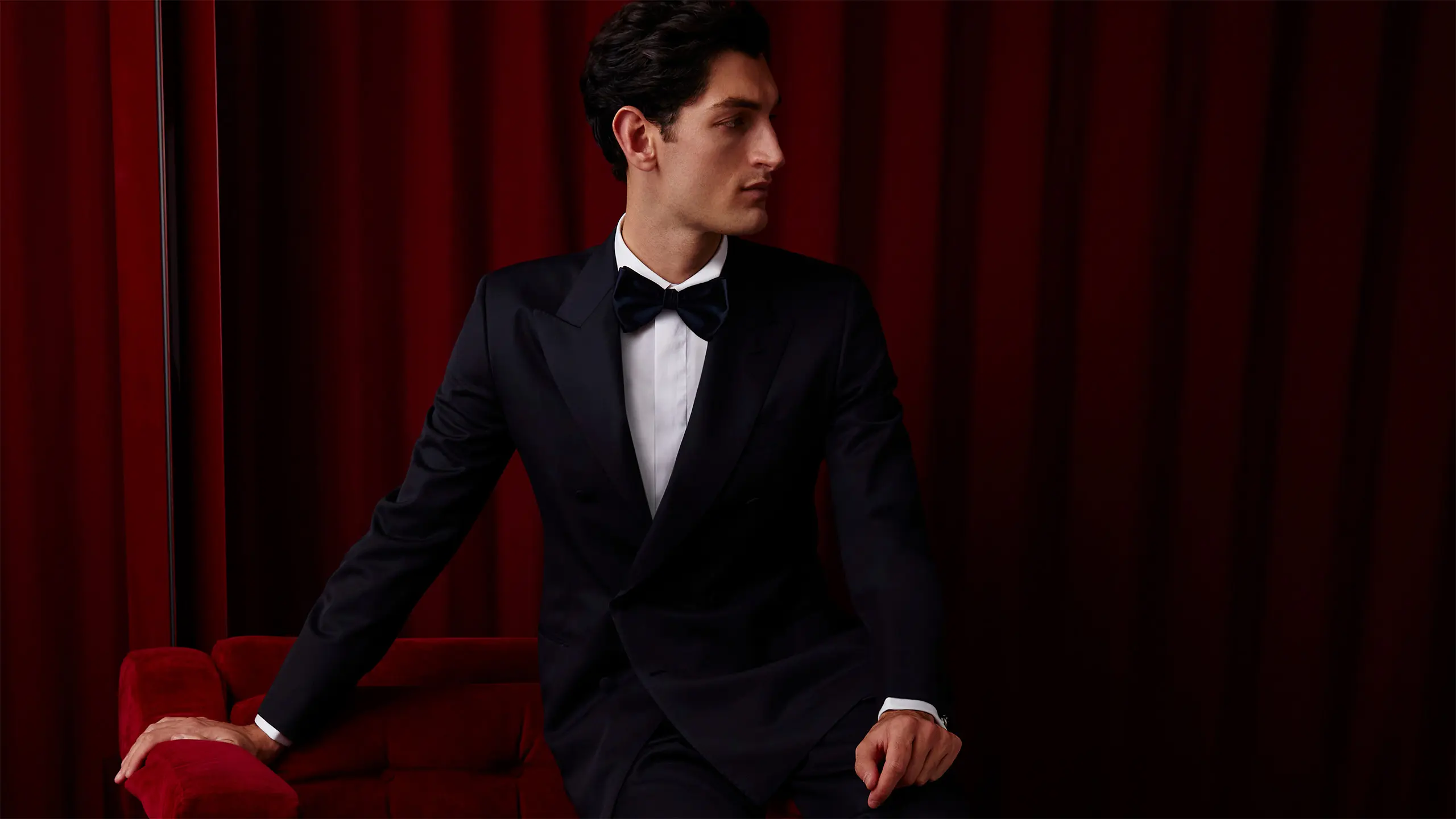 Person in elegant Brioni black tuxedo and bow tie seated against rich red velvet curtain backdrop.