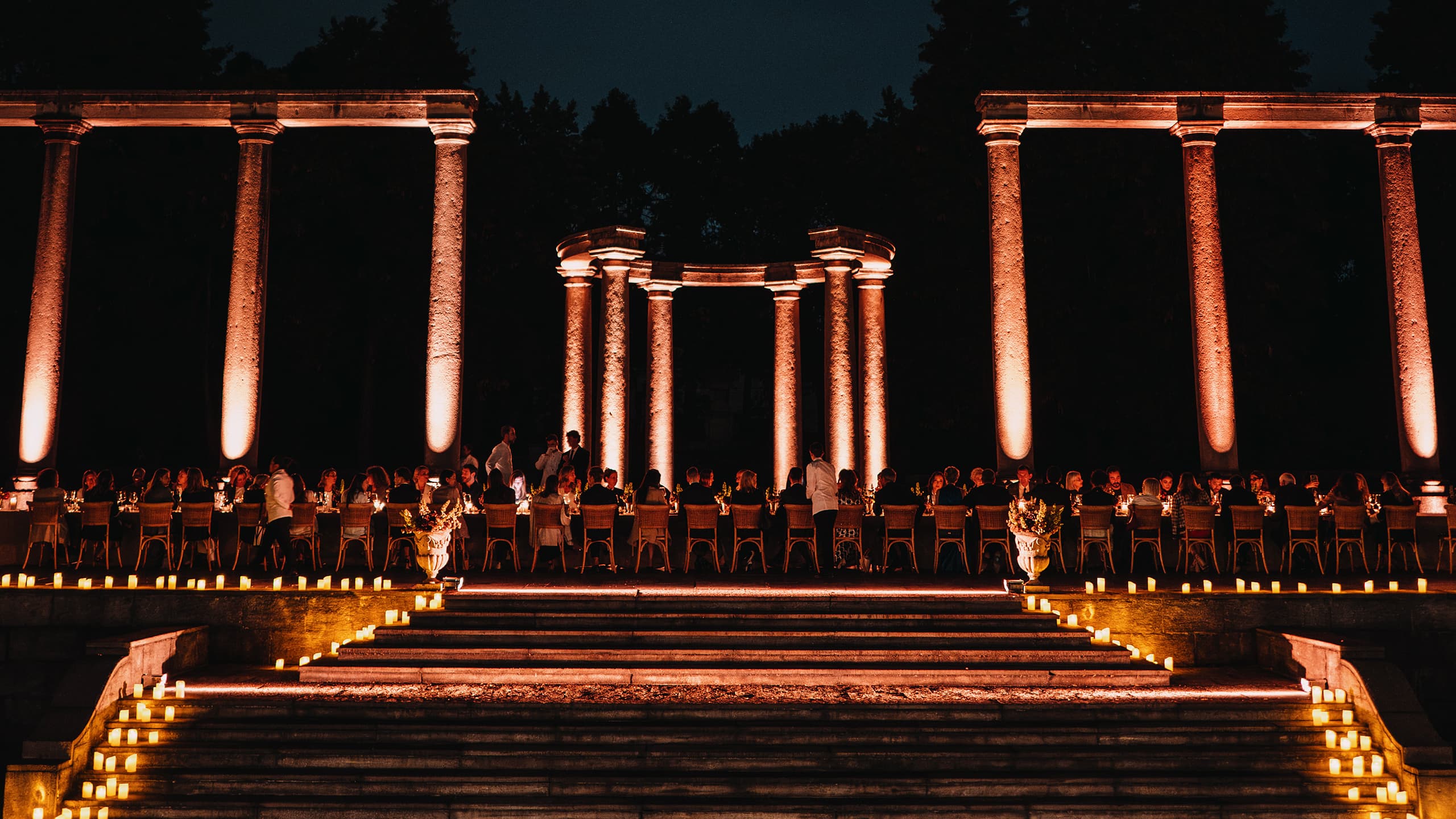 Evening outdoor Brioni dinner event with guests seated at long tables beneath illuminated orange classical columns and candlelit steps.