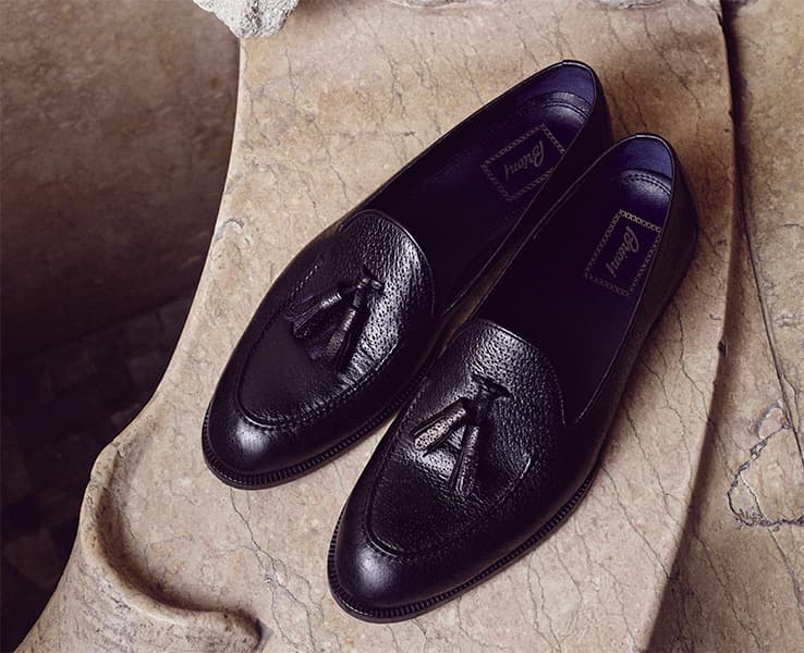 Elegant Brioni black leather tassel loafers displayed on textured beige stone surface.
