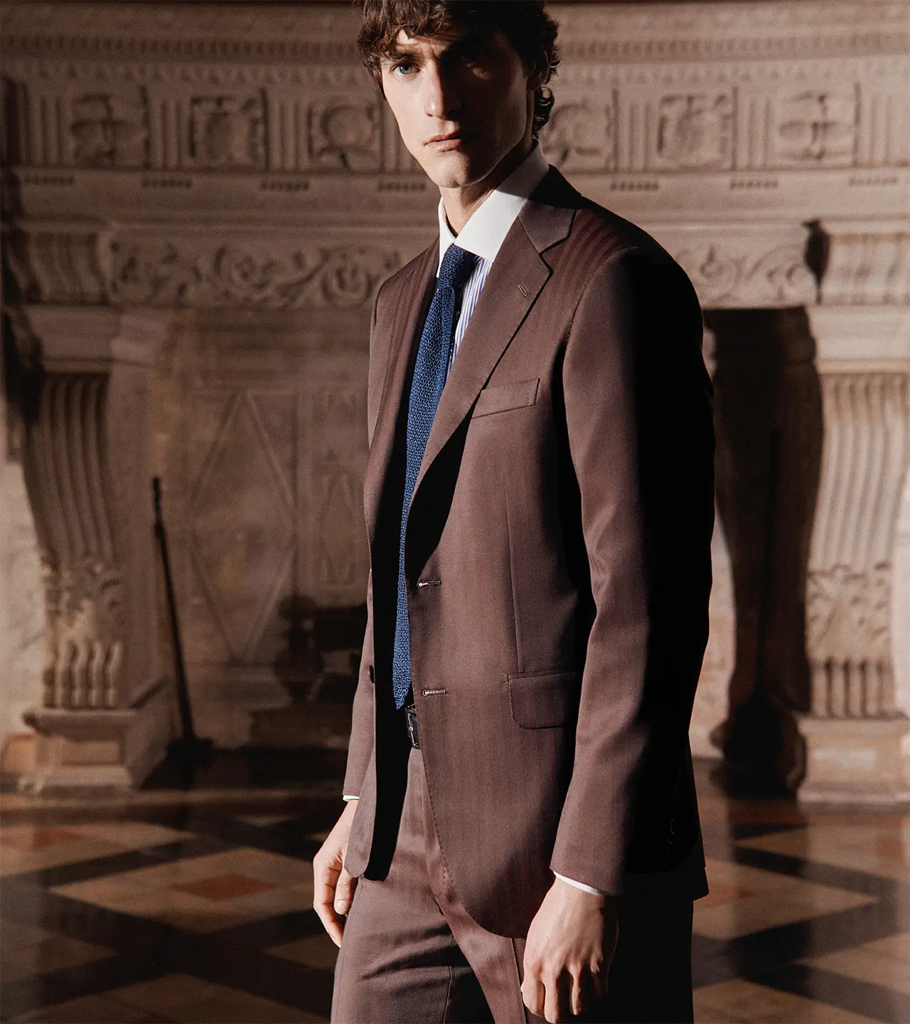 Man in Brioni Iride brown suit with blue tie standing in ornate interior with decorative stone columns and carved architectural details.