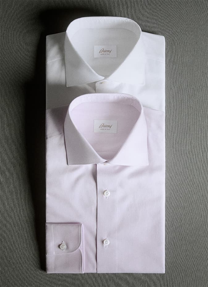 Two folded Brioni formal shirts, one white and one light pink, displayed on a gray textured surface.