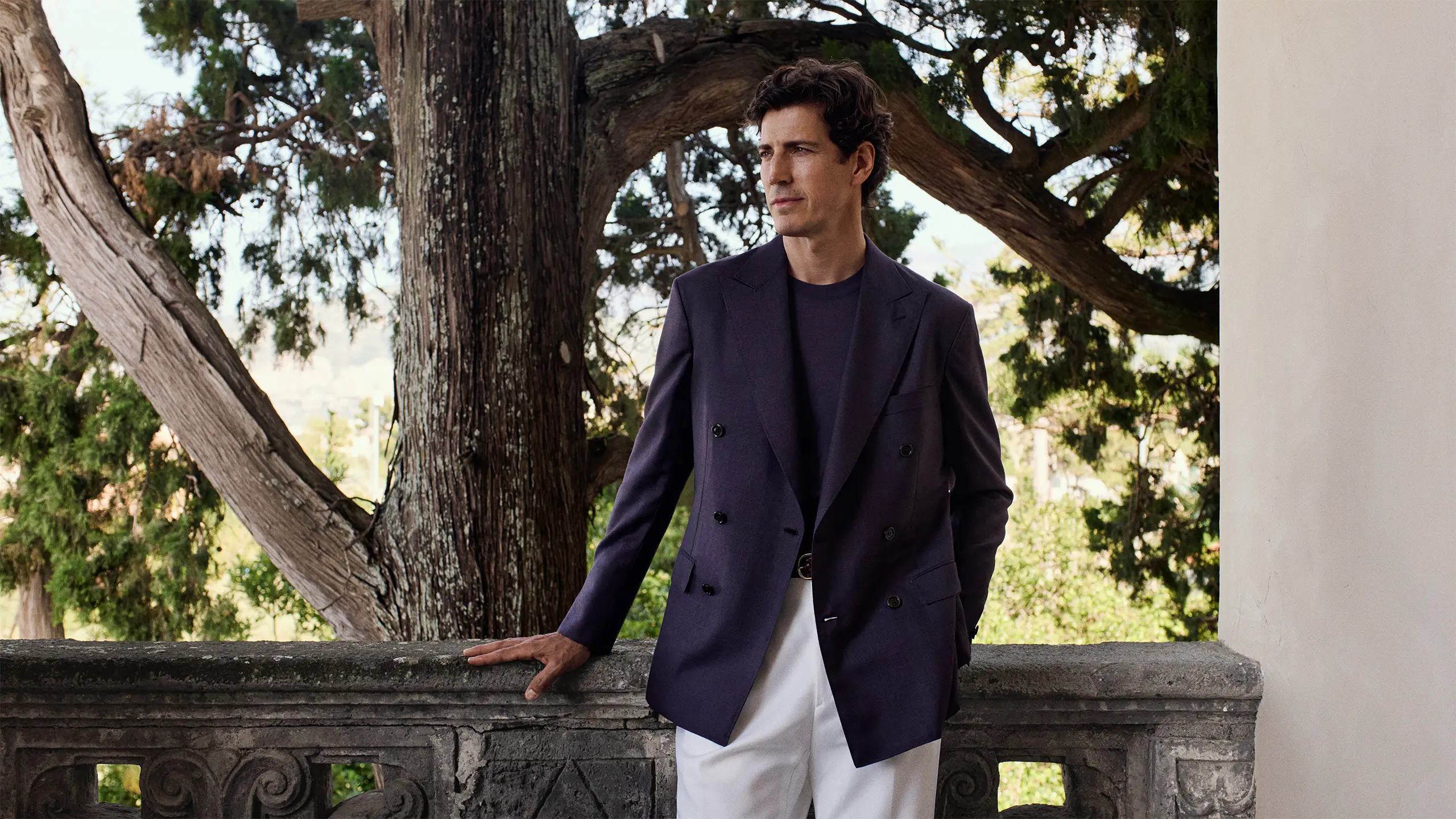 Man in Brioni navy double-breasted blazer and white pants standing by stone balustrade with gnarled tree branches in background.