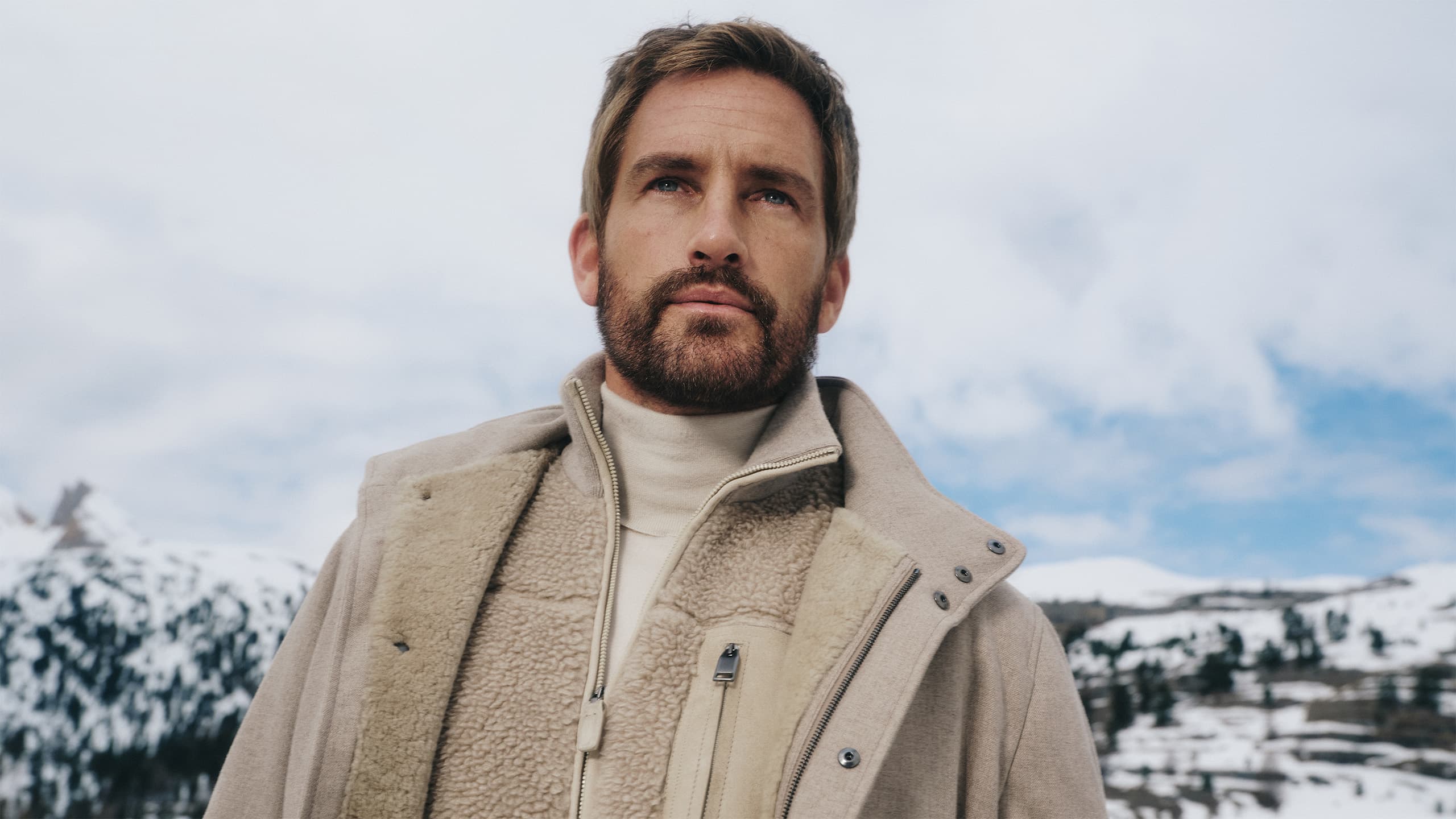 Person in Brioni beige sherpa-lined coat gazing thoughtfully against snowy mountain backdrop under cloudy sky.