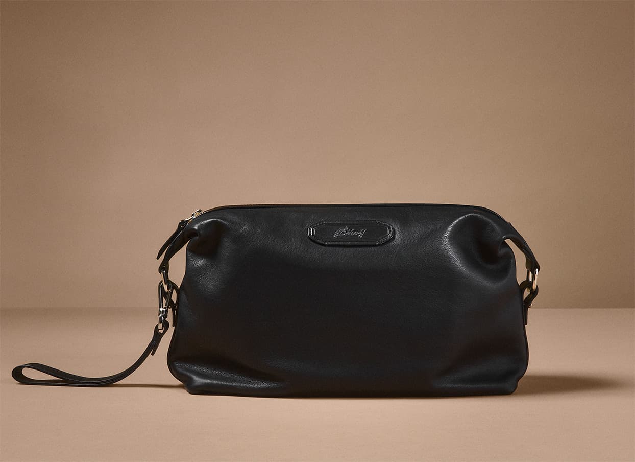 Brioni black leather toiletry bag with wrist strap displayed against a neutral beige background.