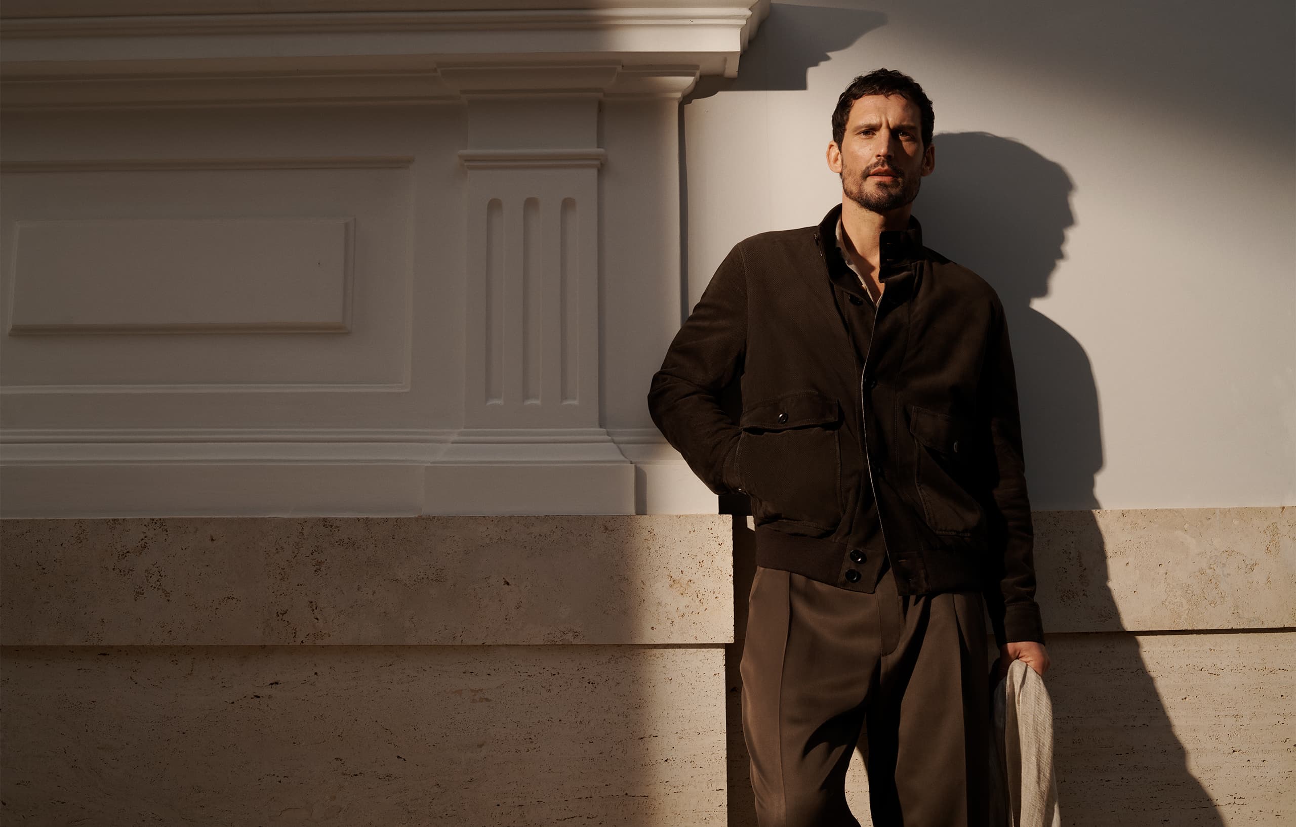 Person in Brioni brown jacket standing against white wall with dramatic shadow, warm golden lighting highlighting elegant attire.