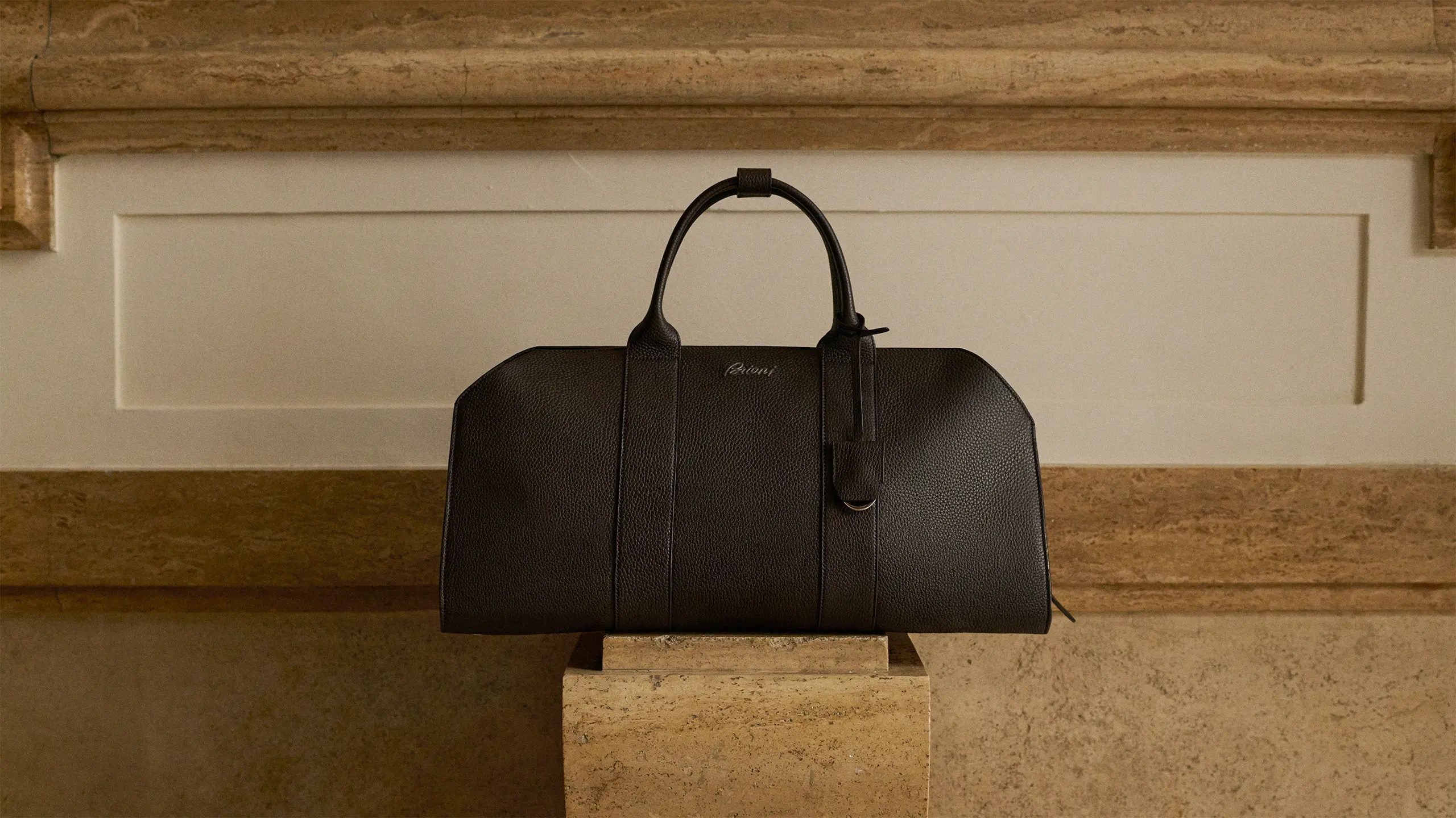 Black leather Brioni duffle bag with handles displayed on stone pedestal against cream cabinet and textured stone wall.