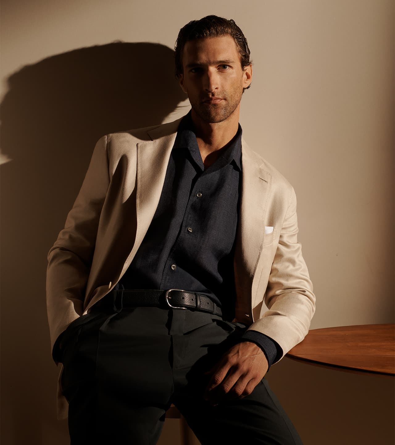 Man in Brioni beige blazer and navy shirt sitting against beige wall with dramatic shadow, wearing black pants and belt.
