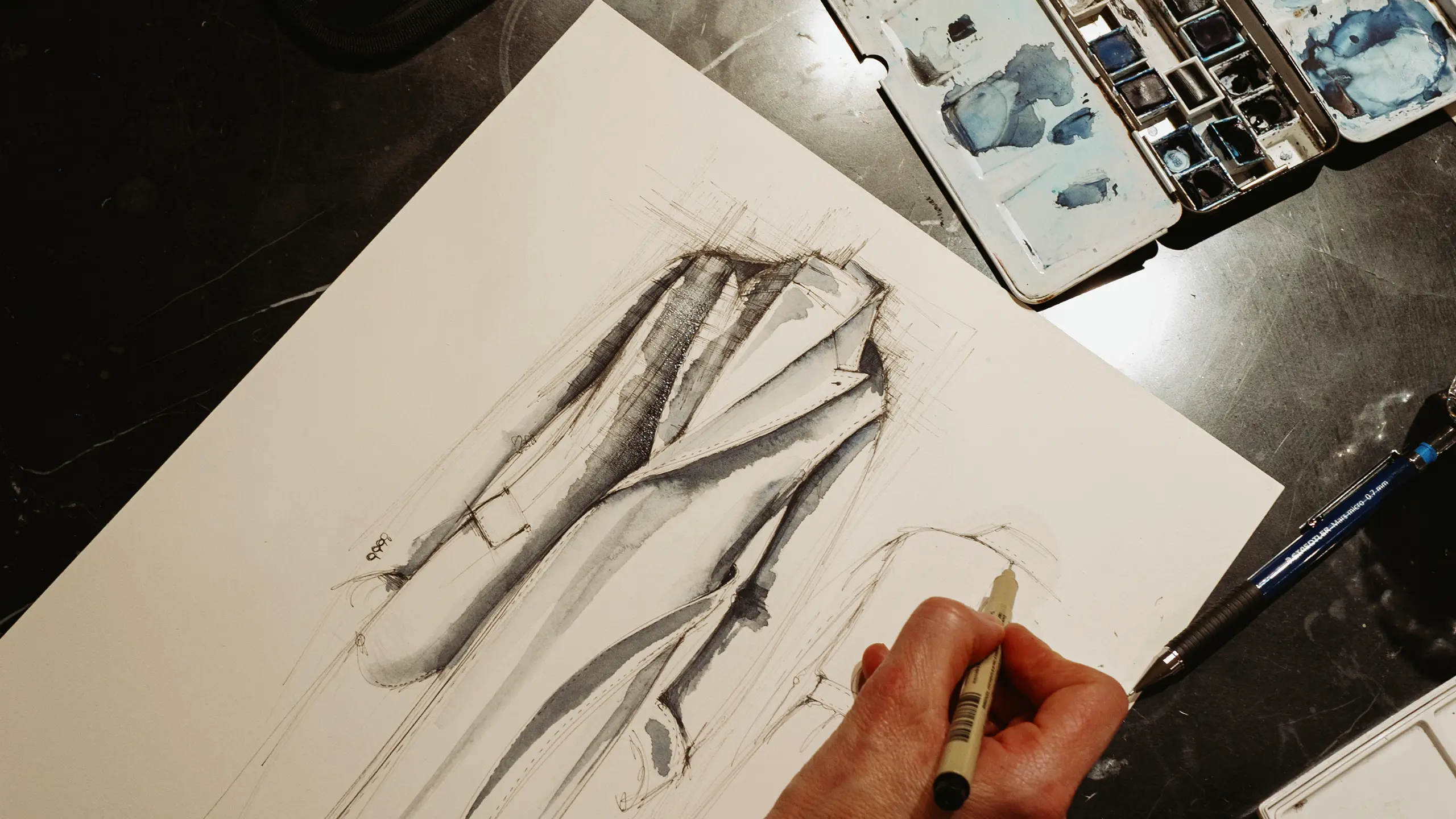 Artist's hand holding brush over watercolor sketch of draped fabric in brown and gray tones, with paint palette nearby.