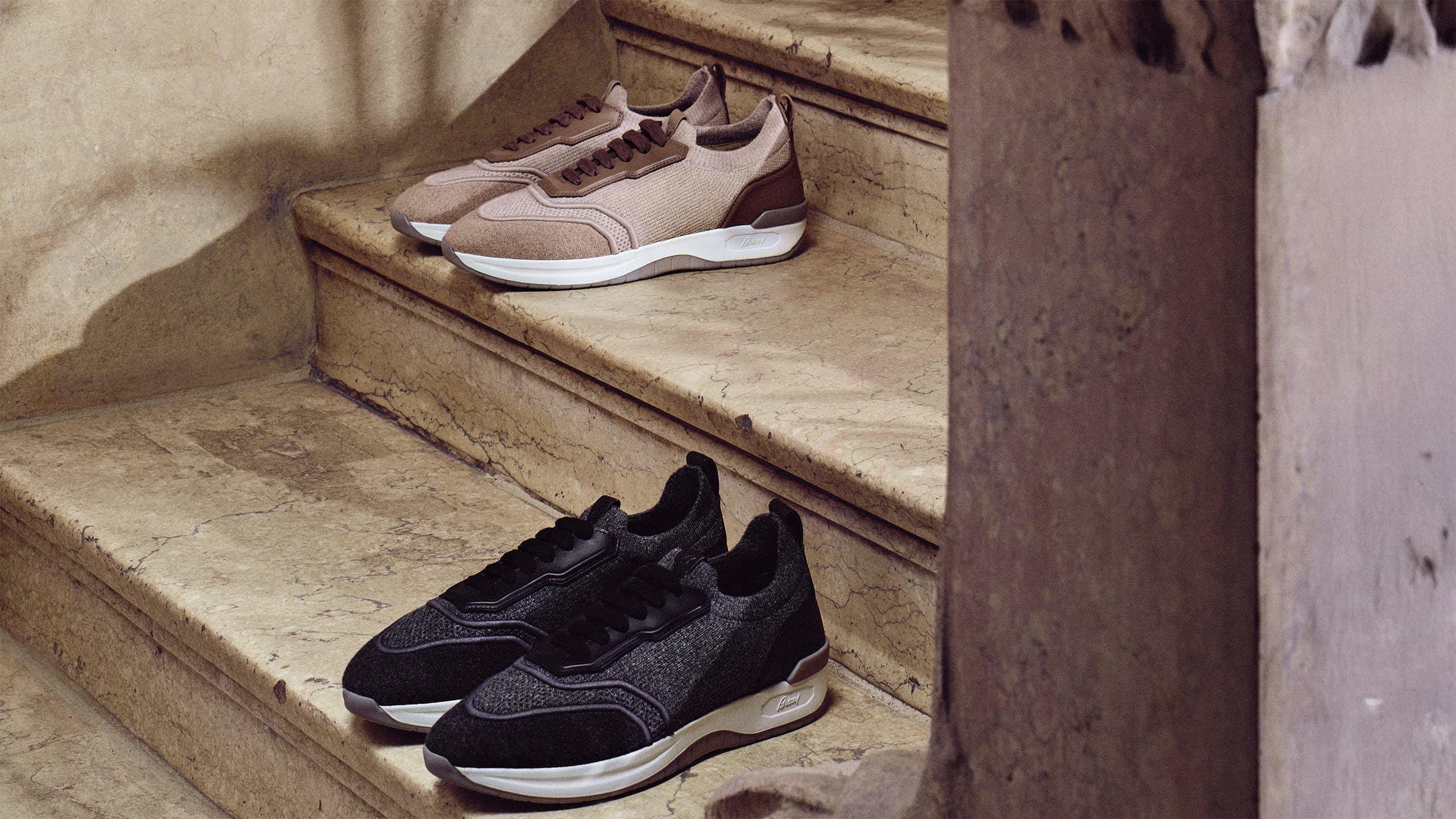 Two pairs of Brioni knit sneakers on stone steps - beige ones on upper step and black ones on lower step.