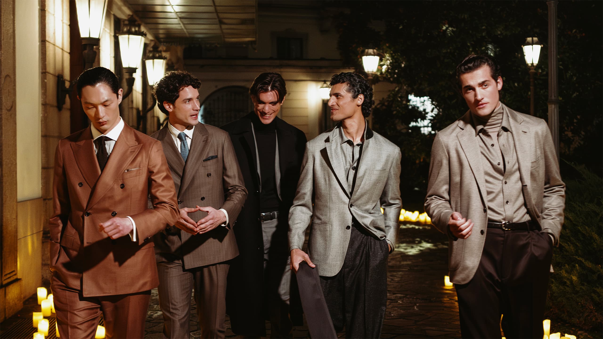 Five men in Brioni elegant suits walking together at night in an illuminated courtyard with candles and warm lighting.