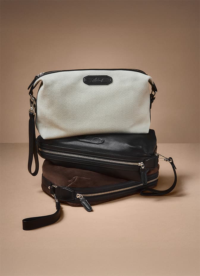Three Brioni travel bags in cream, black, and brown leather with zipper details against a neutral beige background.