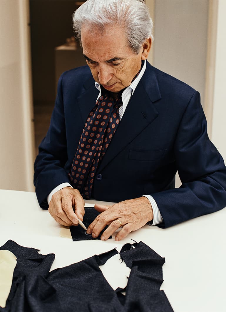 Un anziano sarto in abito blu navy con cravatta a fantasia lavora meticolosamente su pezzi di tessuto scuro a un tavolo bianco.